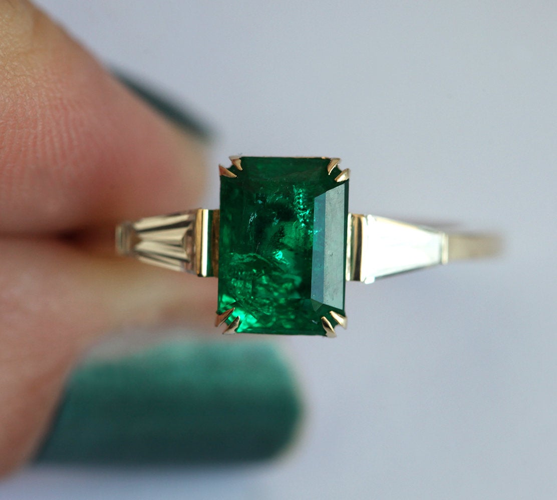 Nahaufnahme eines goldenen Smaragdrings mit Diamantakzenten am Finger, der leuchtend grüne Smaragde und funkelnde Diamanten zeigt. Perfektes Jubiläumsgeschenk für ihn.