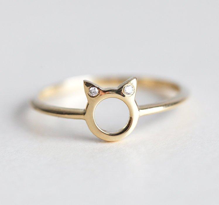 Ein goldener Ring in Form von Katzenohren mit runden weißen Diamanten an den Ohren, auf einem strukturierten Hintergrund