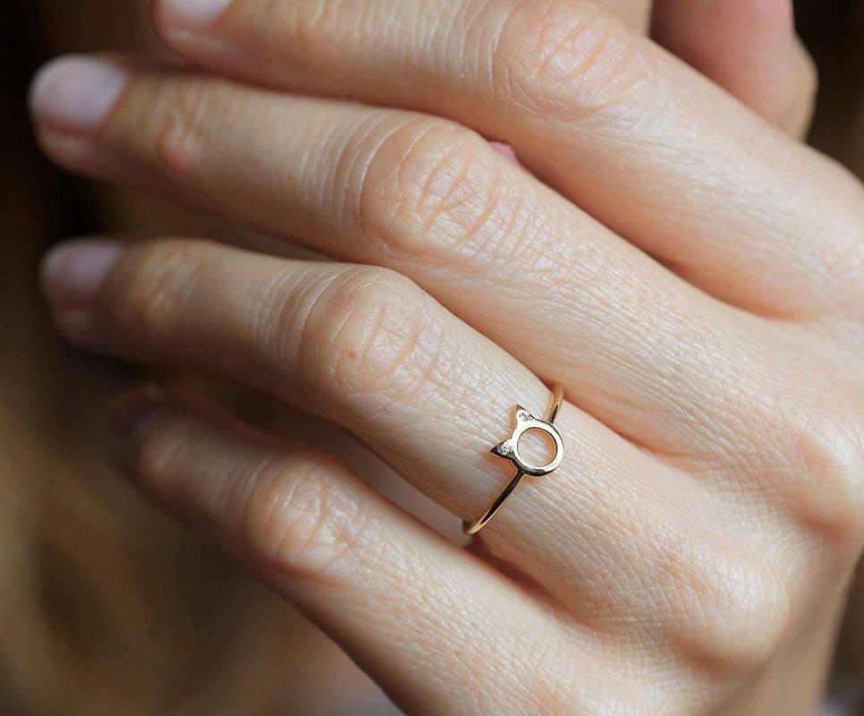 Ein goldener Ring in Form von Katzenohren mit runden weißen Diamanten an den Ohren, auf einem strukturierten Hintergrund