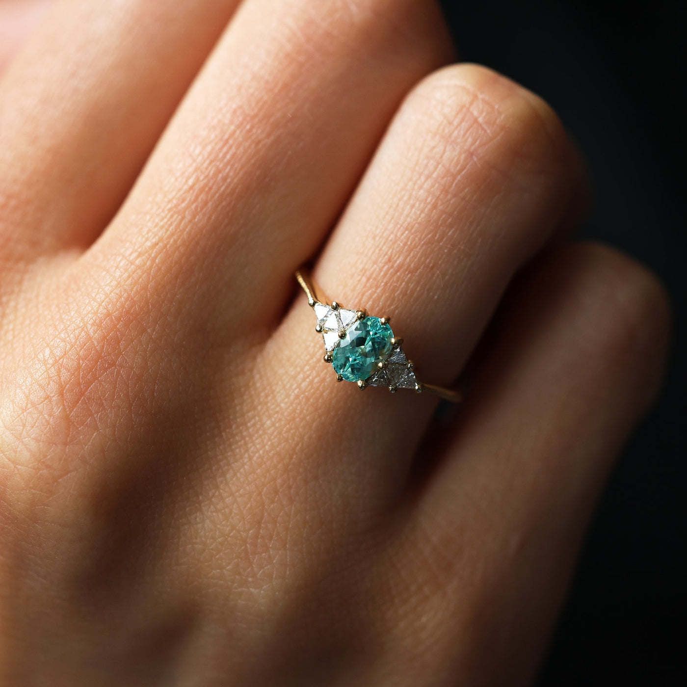 Eine Hand trägt einen Paraiba-Turmalin-Verlobungsring mit Diamant-Seitensteinen. Ovaler Paraiba-Turmalin-Mittelstein mit dreieckigen Diamanten an den Seiten. Hergestellt aus 14-karätigem oder 18-karätigem Gold oder Platin.