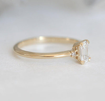 Diamantring im Smaragdschliff mit seitlichen Diamanten auf weißer Oberfläche, der Eleganz und Brillanz zur Schau stellt.