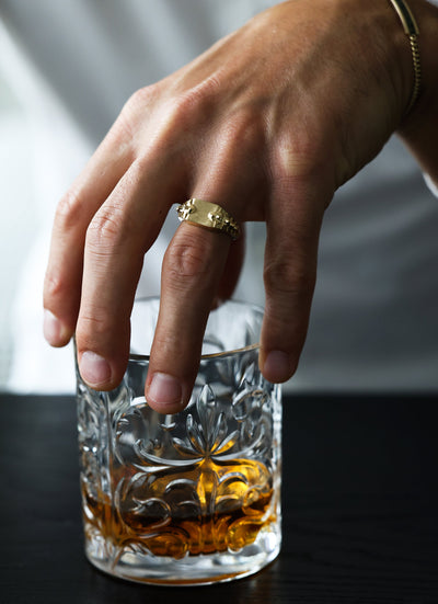 Eine Hand, die einen goldenen Schlangen-Siegelring über einem Glas Alkohol trägt.